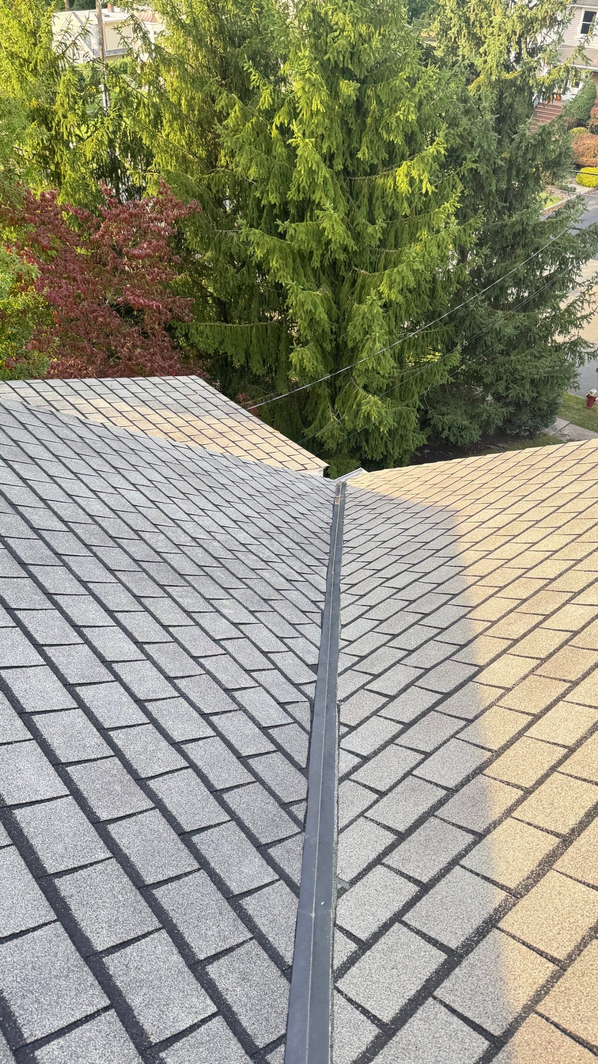 Open metal valley between two weathered shingle roof planes on a Randolph, NJ home, with tall spruce trees beside the house